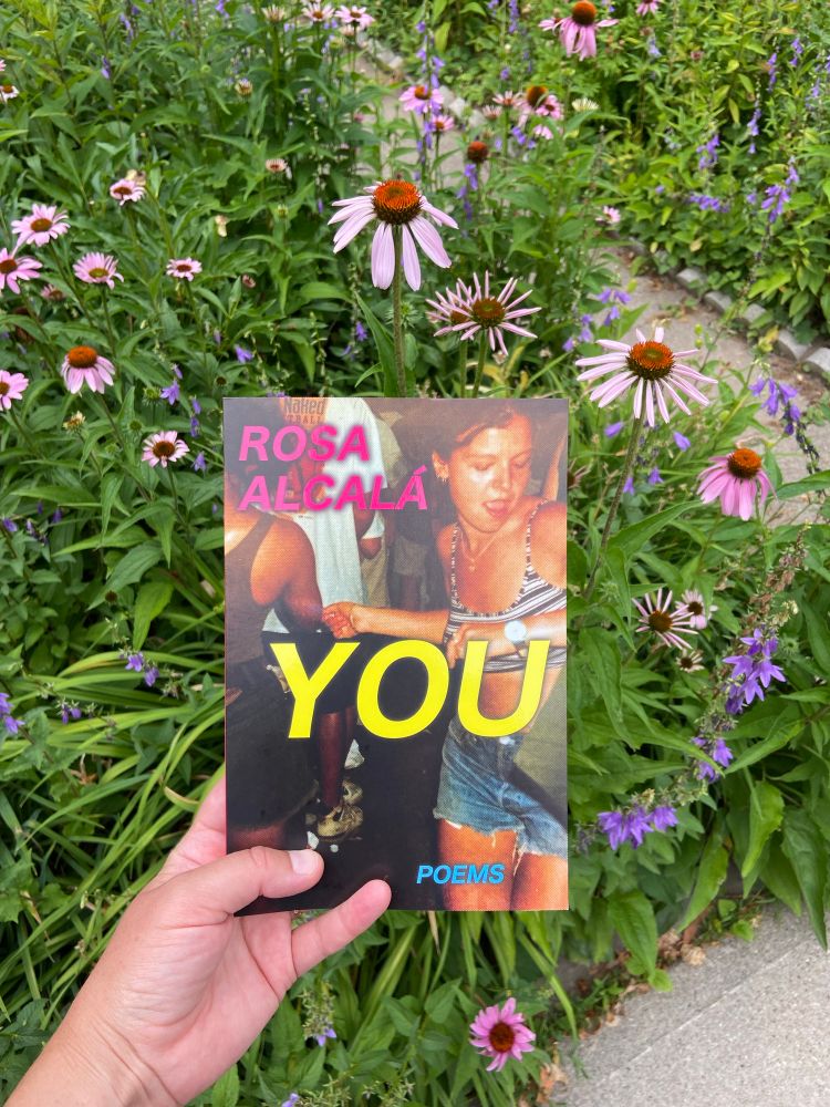 A hand holding Rosa Alcalá’s book YOU, with echinacea flowers in the background 