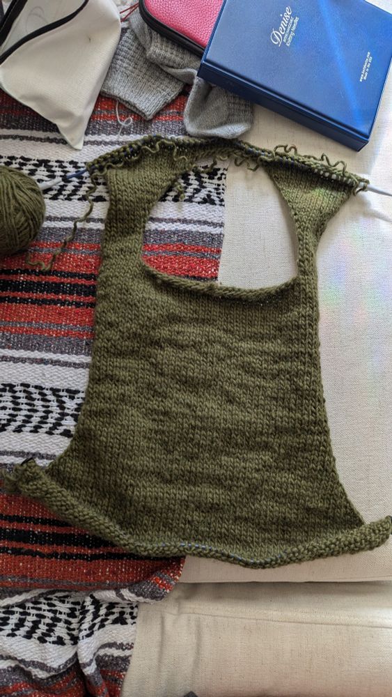 Photo of a half knit olive green vest on a red, white, black and grey woven blanket. Knitted socks and knitting accessories in the back.