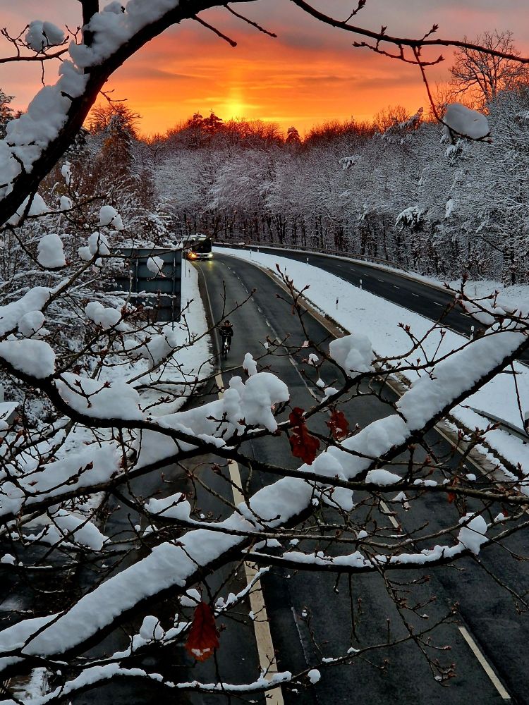 Blick von Brücke auf Straße. Ein Radfahrer und ein Bus sind zu sehen. Umrandet von Schnee. Roter Sonnenuntergang im Hintergrund.