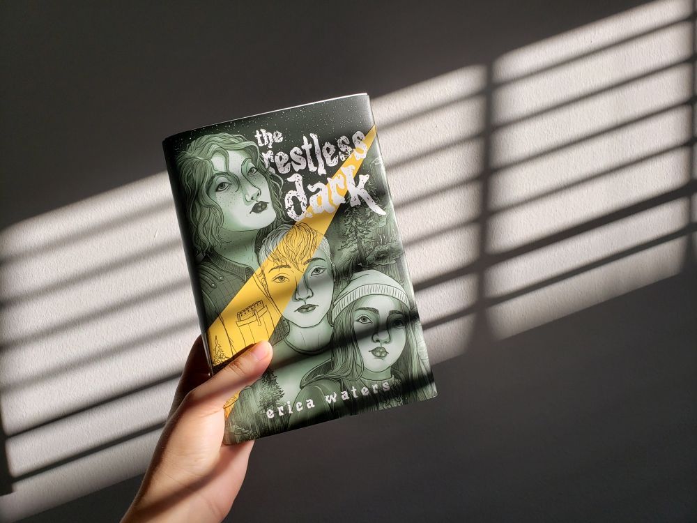 Photograph of THE RESTLESS DARK Rainbow Crate special edition, in dramatic blinds lighting. The cover shows three girls' busts in a green monochrome with a steak of yellow light across the cover.
