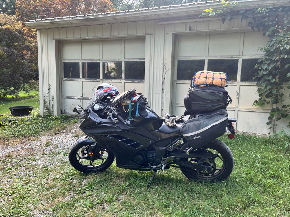 A photo of a black Kawasaki Ninja 300 heavily loaded for touring, parked in front of a weathered garage.
