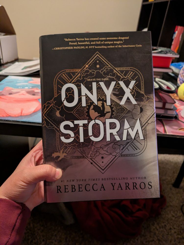 Person holds a book that reads "Onyx Storm from Rebecca Yarros" the book cover is mainly black and grey with outlines of gold