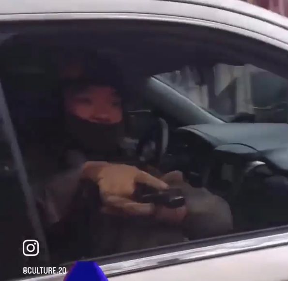 Masked ICE thug sitting in a car with an open window, aiming a handgun at the bystanders in the crowd.