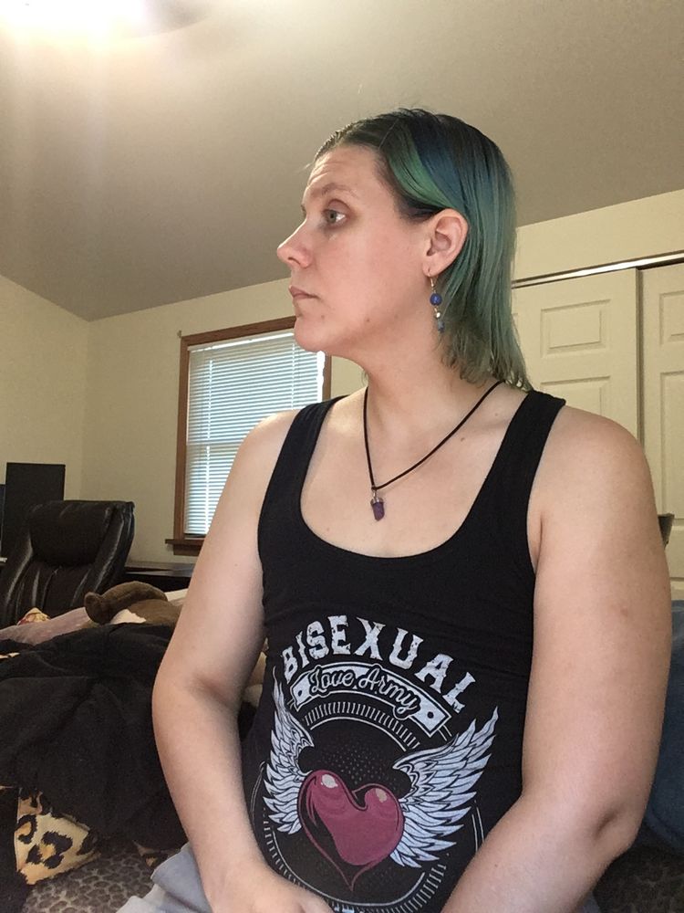 white nonbinary person, blue hair, earrings and a necklace, wearing a tank top saying "Bisexual Love Army" with wings around a heart.