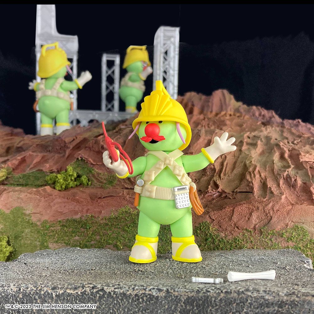 The Fraggle Rock Flange Doozer figure , a small green fellow in a construction hard hat, radioing instructions to other Doozers on a faux-rocky backdrop.