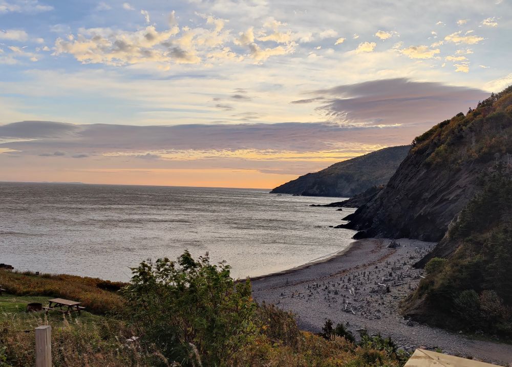 The view from my patio: looking out over Meat Cove Beach (dotted with cairns and bits of wood) with sloping rock faces. Pale orange of sunrise glowing from behind the furthest outcrop.