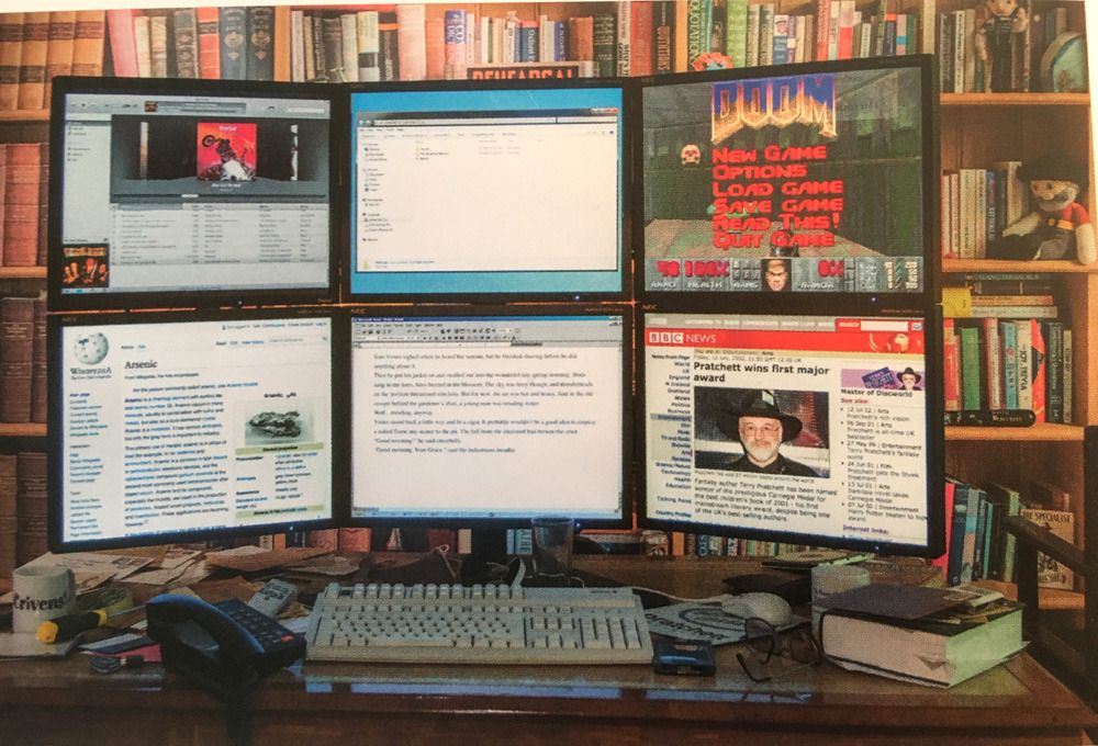 A picture of Terry Pratchett's 6 monitor setup, in front of a wall of books. Notably on the screens is Wikipedia, iTunes, Doom, and a BBC article about Terry Pratchett. Each monitor is enormous. 