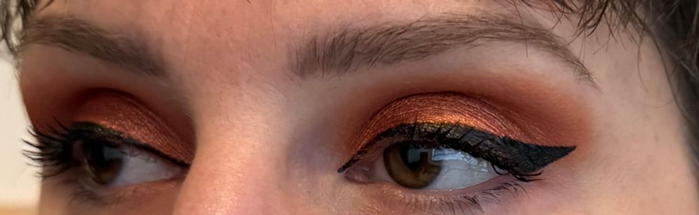 A close-up of Wanda’s eyes. They’ve used a fiery copper eyeshadow on their entire lid, buffed out using a warm orange-brown, and their eyeliner is a thick cat’s eye shape. In this photo, they are looking to their right, with their left eye in the light and the right in shadow.
