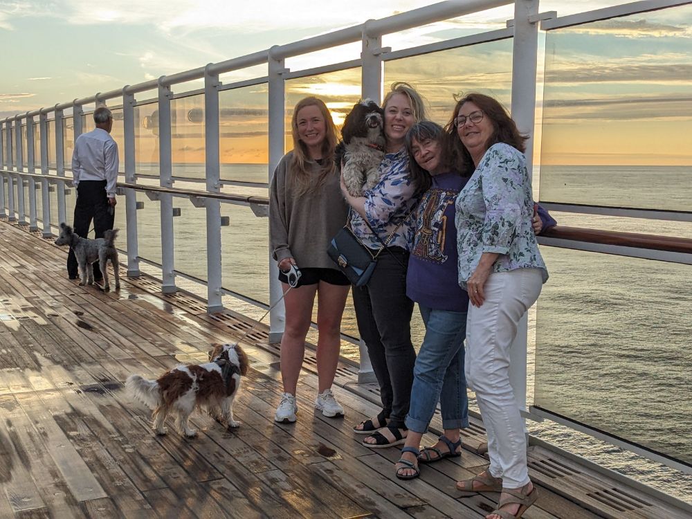 In the foreground, 4 women and 2 dogs, huddle close for a photo. In the background is a man with his black and white dog gazing out on the setting sun. 
