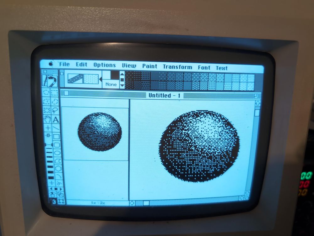 Picture of SuperPaint running from a Macintosh Plus, the blue hue of the CRT shows SuperPaint opened and a graphic of a shaded 3d ball is being worked on. A zoomed out and zoomed in view are side by side on screen. We can see the classic user interface with paint tools that were established in this era on the left side, like paintbrush, lasso select, eraser, etc. 