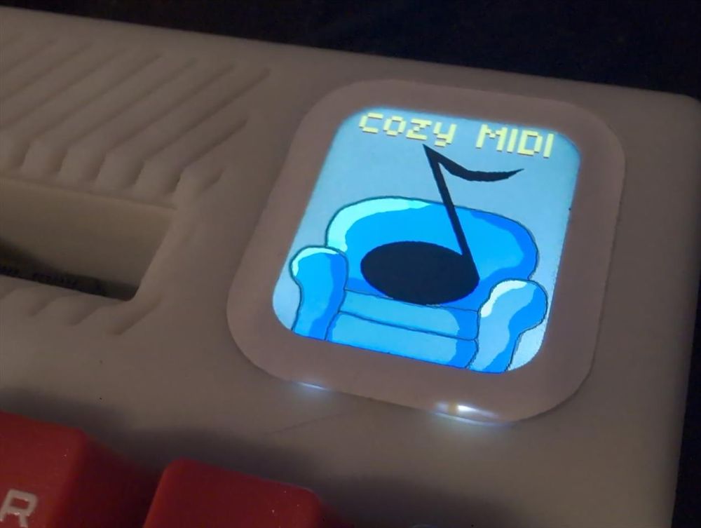 a watch sized LCD screen embedded into the F256K2 case, showing a logo of the program cozy MIDI. it's a cartoon 1 person couch in blue, upon which a big black note is sitting. The words 'cozy MIDI' are shown at the top in yellow.