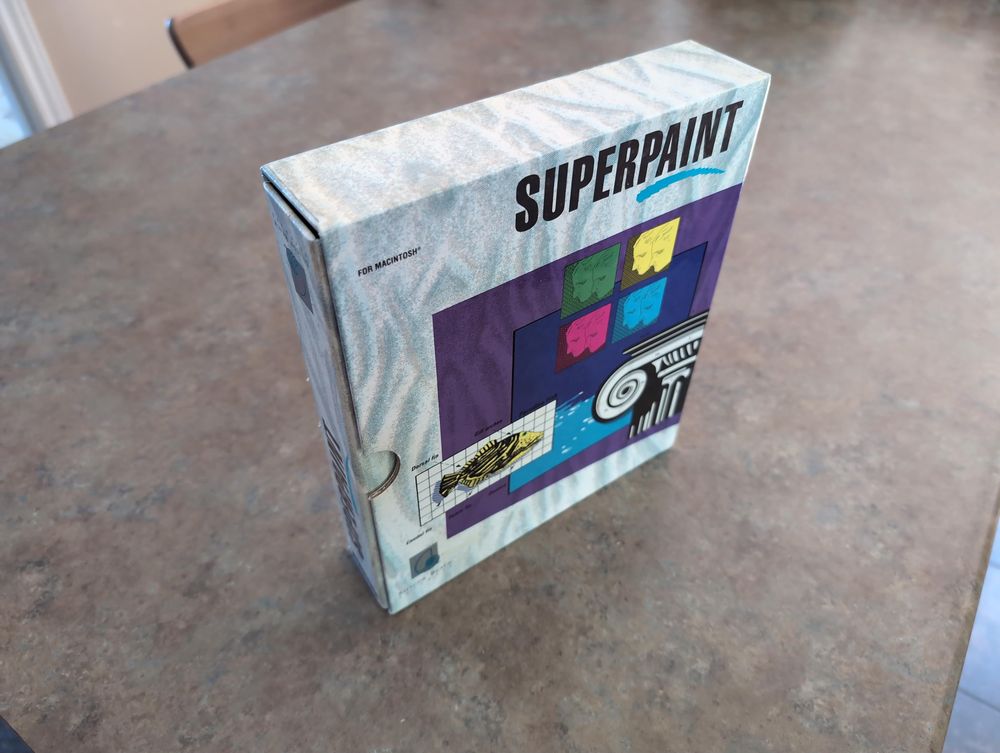 Top down angled view of SuperPaint's software box showing a definitely 80's pastel design on the front. We can see a ring binder's side that can slide out of the box frame.