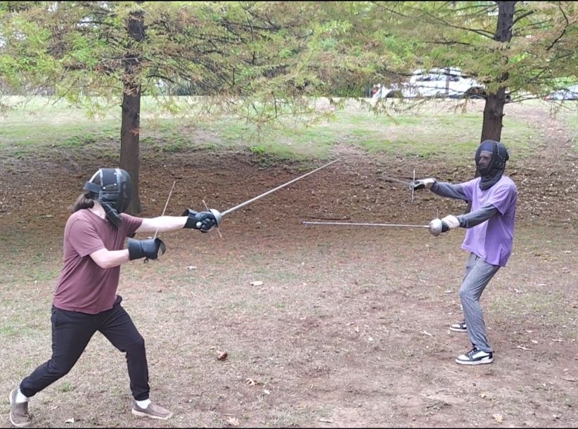 Two men dressed in light workout clothes fencing masks and heavy gloves facing each other and wielding rapier and parrying daggers.