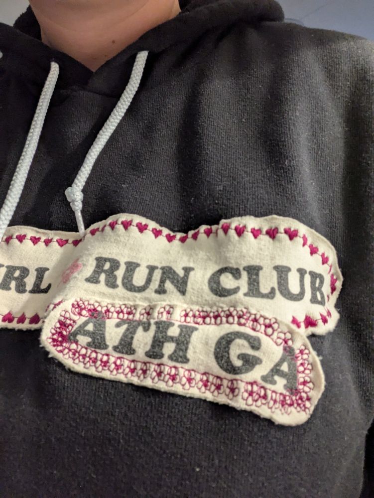 closeup of a black hoodie with patches cut out from a cream hoodie reading "slow girl run club, ath ga" on it. they're stitched on with bright pink thread in hearts and flowers
