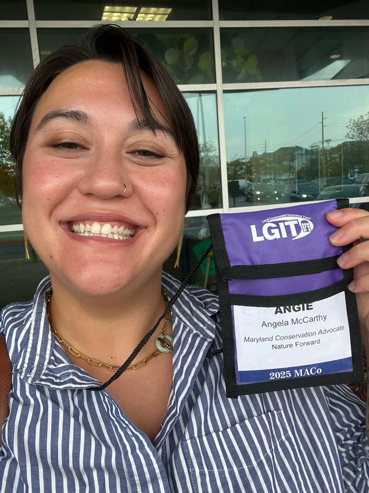 Angie holding up a MACO badge with a big smile on her face. She’s wearing fish earrings and is wearing a blue and white striped shirt. 