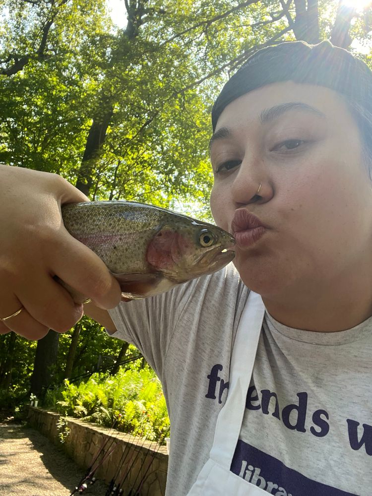 Angie kissing a rainbow trout at the Becoming an Outdoorswoman weekend sponsored by MD DNR. Her hat is backwards and she’s wearing a Mount Pleasant library t shirt. 