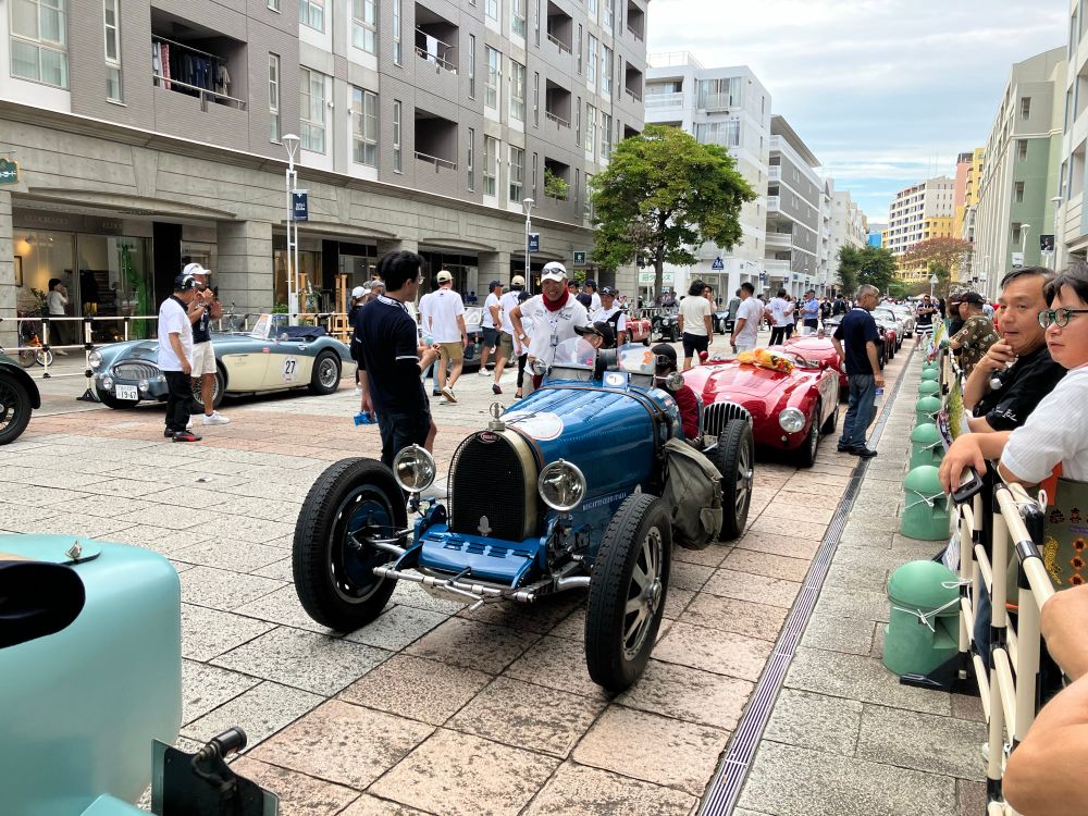 Two rows of 20th century cars lined up along the Baytown Promenade. Dozens of people gathered along the barricades watch as driving team members mingle among the vehicles. 