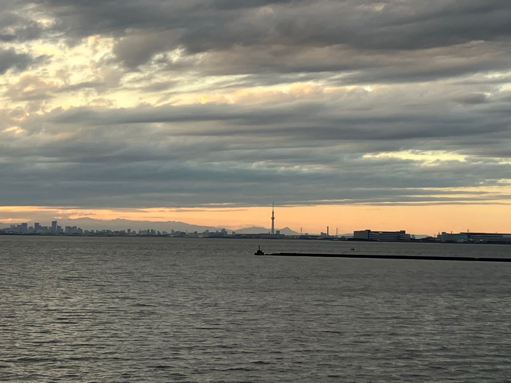 Layers of clouds above an orange sky. Tokyo SkyTree is visible in the center distance. Tokyo’s skyline is silhouetted to the left. Tokyo Bay is in the foreground. 