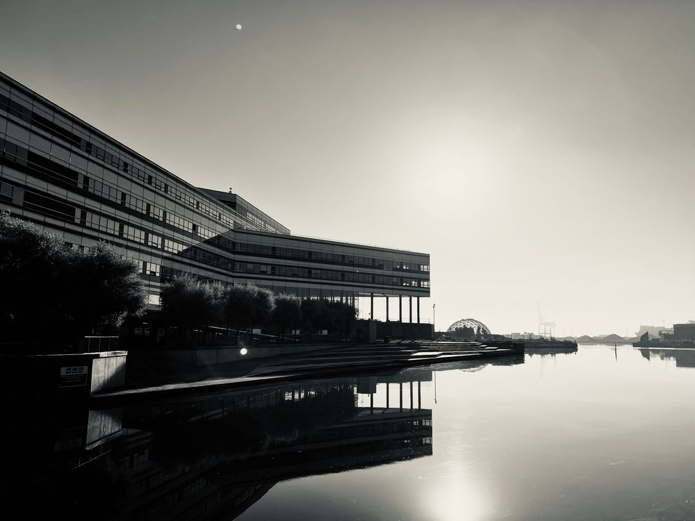 Moderne Gebäude im alten Stadthafen von Aarhus spiegeln sich im Wasser. Im Hintergrund, nur als Silhouette zu erkennen, eine große Kranbrücke. Monochrome. © Torsten Grieger