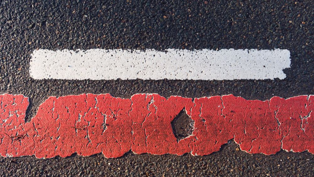 Trennstreifen zwischen einem viel zu schmalen Radweg und einer viel befahrenen innerstädtischen Straße in Altenburg. Im Hintergrund schwarzer Asphalt, darauf eine schmaler weiße Trennlinie und Reste der roten Radweg-Markierung.
