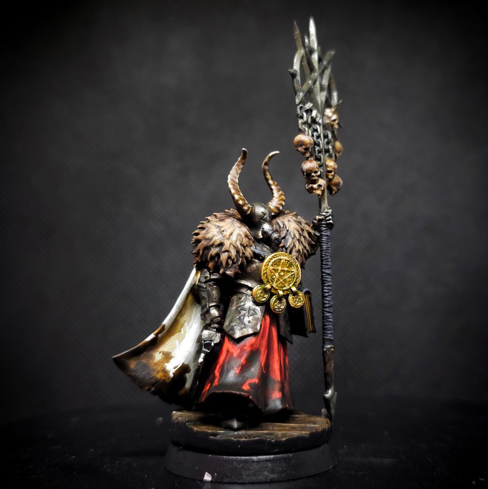 Photo of a painted miniature for Trench Crusade; a Heretic Legions Heretical Priest. A humanoid in heavy, tarnished plate armor and muddy red robes. They wear a filthy white cloak and brown fur over their shoulders. They have a glossy Obsidian Sickle in one hand and with the other they heft a staff tipped with a pentagram and several human skulls. A weighty tome hangs from their hip and a Blasphemous amulet hangs around their neck. They wear a horned helmet with a gas mask filter.