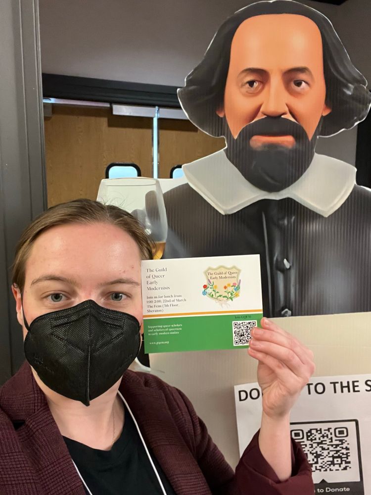 A woman in a red blazer and black mask stands next to a cardboard cutout of Shakespeare at the Shakespeare Association of America. She is holding a postcard for The Guild of Queer Early Modernists 