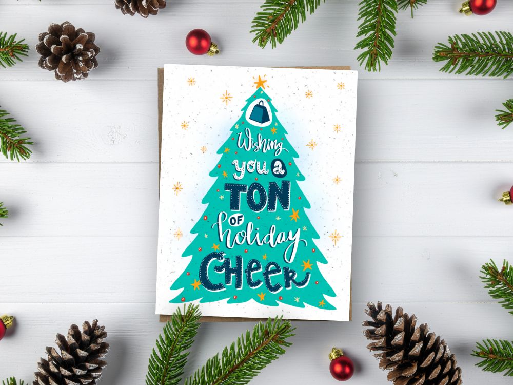 physical greeting card on a holiday themed table. the card has a graphic of a christmas tree and says "Wishing you a TON of holiday cheer"