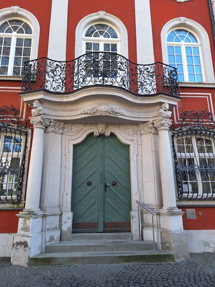 Ein Portal an einem Gutshaus oder Schloss, das rot gestrichen ist. Das Portal ist mit Säulen verziert und endet oben in einem Balkon, der über dem Portal ist. Die Tür ist grün gestrichen. 