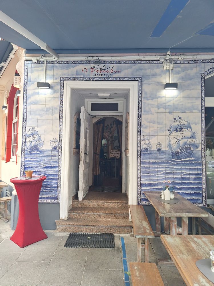 Ein einfacher weißer Türrahmen, der in einen kleinen Vorraum führt und dann eine weitere geöffnete weiße Tür, der ins Innere des Hauses Führt. Die Außenfassade ringsum die Tür besteht aus Fließen, die in blau und weiß gehalten sind und das Meer mit Segelschiffen zwigen. 
Vor dem Gebäude zwei Tische mit Bänken aus Holz und ein Stehtisch mit einer roten Husse.