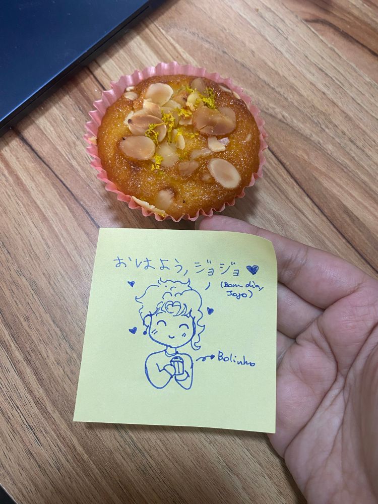 Picture of a hand resting against the wooden table top, holding a green sticky note that has a little drawing of Kakyoin from Jojos holding a tiny cake. It written “good morning jojo” in both Japanese and Brazilian Portuguese. A little part of our laptop is showing at the top left corner of the picture.