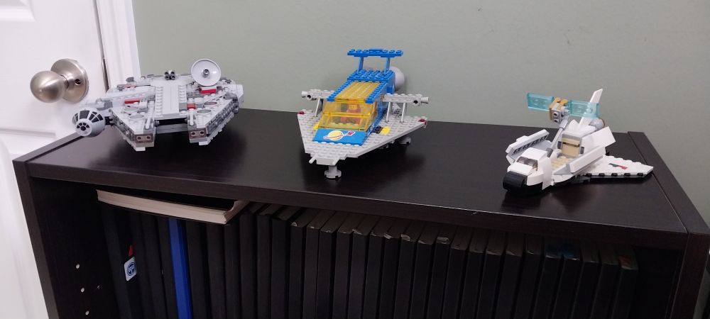 LEGO Millennium Falcon, Space Cruiser, and Space Shuttle from left to right on top of small bookshelf.