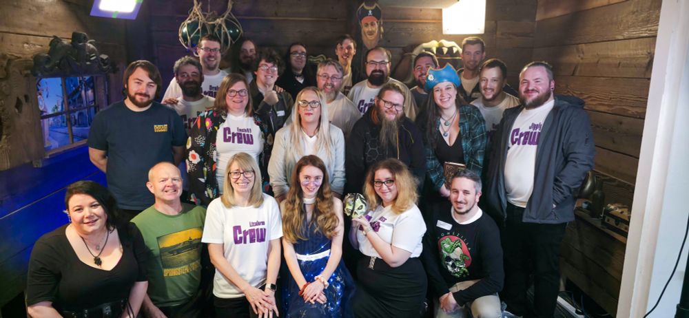 A group of people stands in front of a tavern set smiling. Several are wearing white shirts with purple lettering reading "crew".