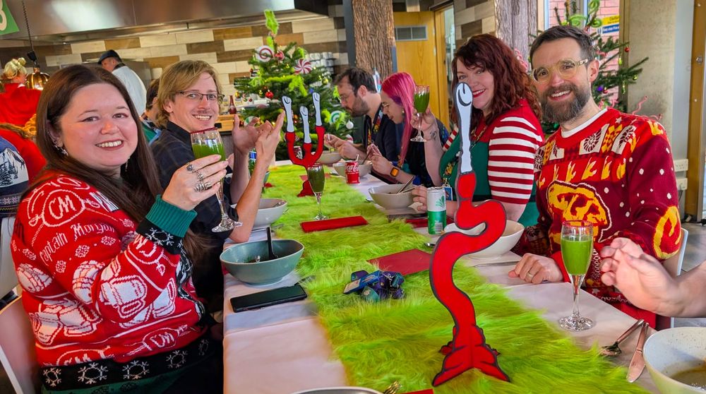 Rare staff look very happy in their overly festive attire. The table they're sitting at is decorated in a long, green, fuzzy tablemat and wiggly red (fake) candles to give the image the air of Seussian whimsy necessary to justify the Grinch's presence.