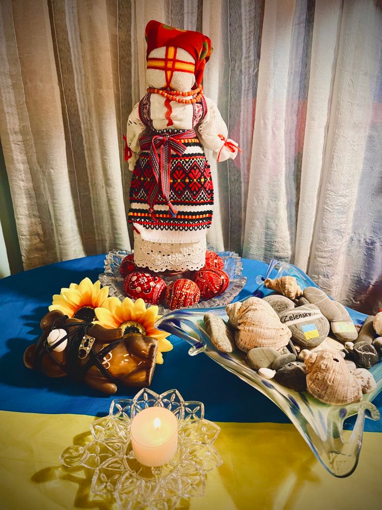 Photo of an altar in support of Ukraine. Altar cloth is the Ukrainian flag. A tealight burns on a glass sunflower candle holder. A square, blue, glass dish holds stones and sea shells from the Black Sea. Two stones are painted. One says, Zelensky and has a Ukrainian flag painted on it. The other says Kyiv. A Russian bear figurine is tied up with rope and a padlock and is lying on its side on a bed of sunflowers. A crystal dish is filled with red pysanky. Above the eggs stands a motanka. Her dress, facial embroidery, and head scarf are red, black, white and orange.