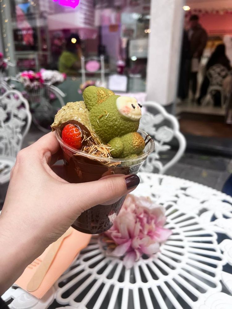 a photo of my hand holding a plastic glass filled with fresh strawberries, belgian hot chocolate,  pistachio paste and a green matcha chocolate labubu on top of it all
