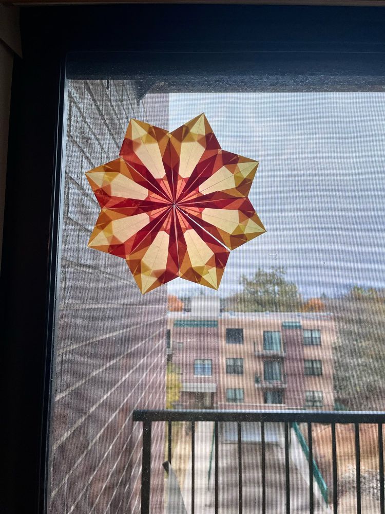 A glass door to a small apartments deck, with another building within our apartment complex across the street. My eight pointed paper star is on the upper left hand corner of photo. The paper star is semi transparent red/yellow gradient.
