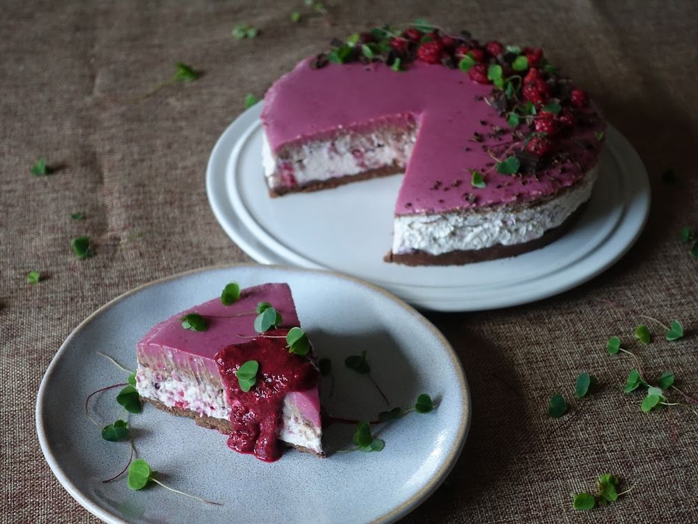 a pink and white cheesecake with a chocolate base decorated with raspberries and green leaves, one slice taken from the cheesecake and displayed on a plate in front of it with a spoonful of raspberry sauce