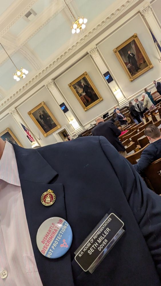 "Dismantle Discrimination, Not Protections" button on my lapel, along with the NH House security pin and my name tag. 

I'm standing on the NH House floor as session is about to begin. 