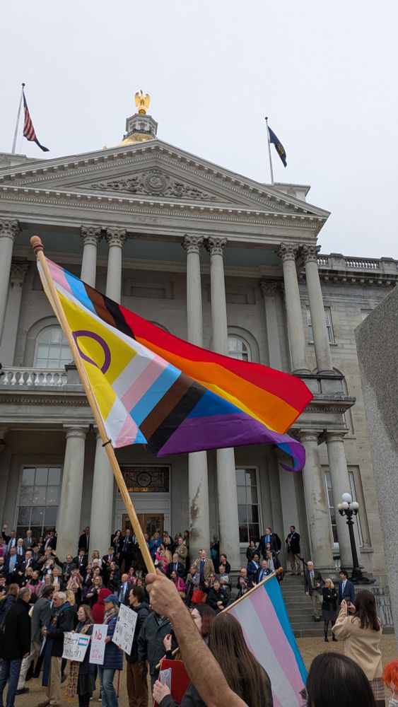 Pride flag waving in front of a crowd standing in front of the NH State House. 