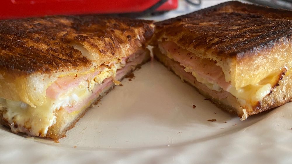 A toasted sandwich cut in half horizontally, showing the sandwich fillings. The sandwich contains mustard, ham, melted cheddar cheese, and a fried egg.