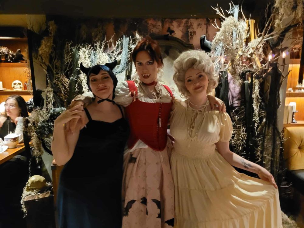 Three lovely ladies pose along a spooky wall. The one on the left is a devil in a blue dress with a thin moustache, the one in the middle is me as a vampire in red and white, and the one on the right is a ghostly Marie Antoinette 