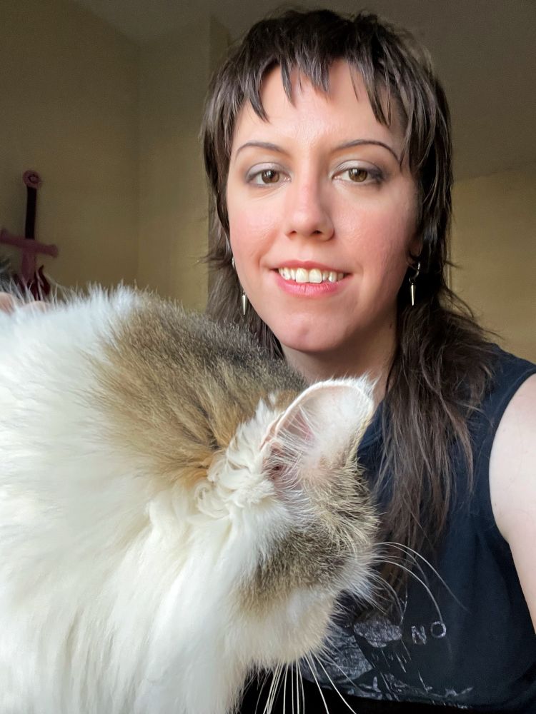 Myself, a feminine person with long brown hair, smiling into the camera. Honey, the most beautiful cat in the world, is in the foreground, her white and great hair so very fluffy and soft.