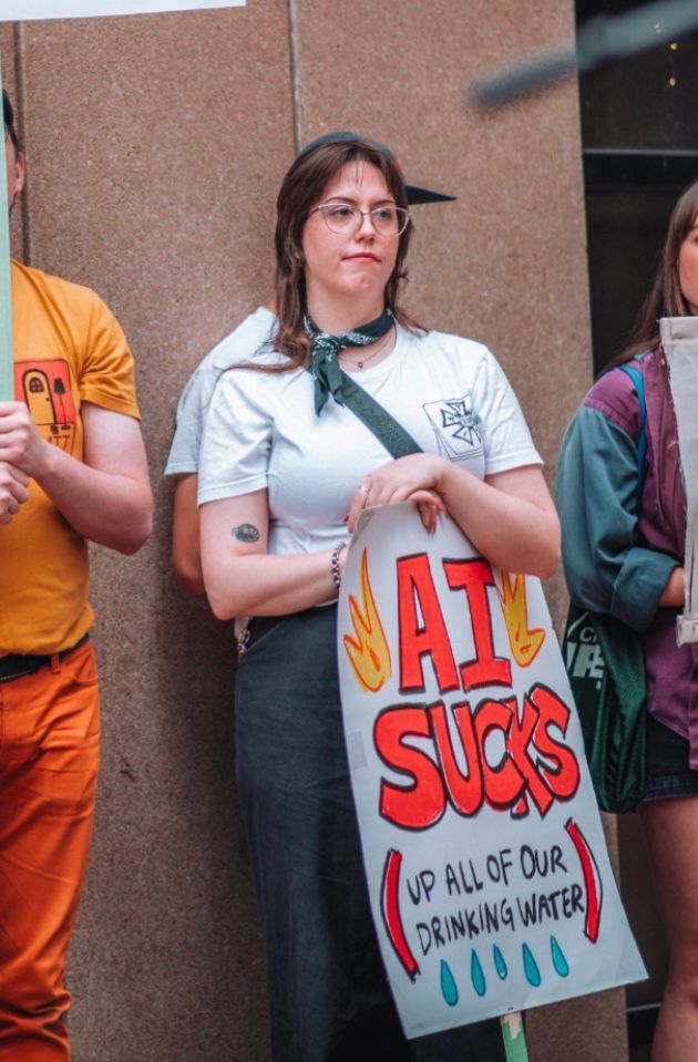 A candid photo of me, standing in a relaxed pose, holding a sign that says “AI SUCKS (up all of our drinking water”
