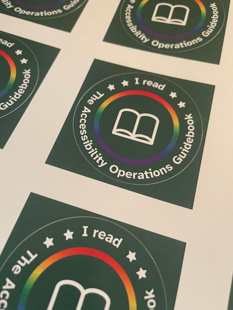 A close-up photo of a sheet of stickers. The stickers are round and dark green with white text that reads 'I read The Accessibility Operations Guidebook' that circles around a rainbow border and a simple icon of a book.