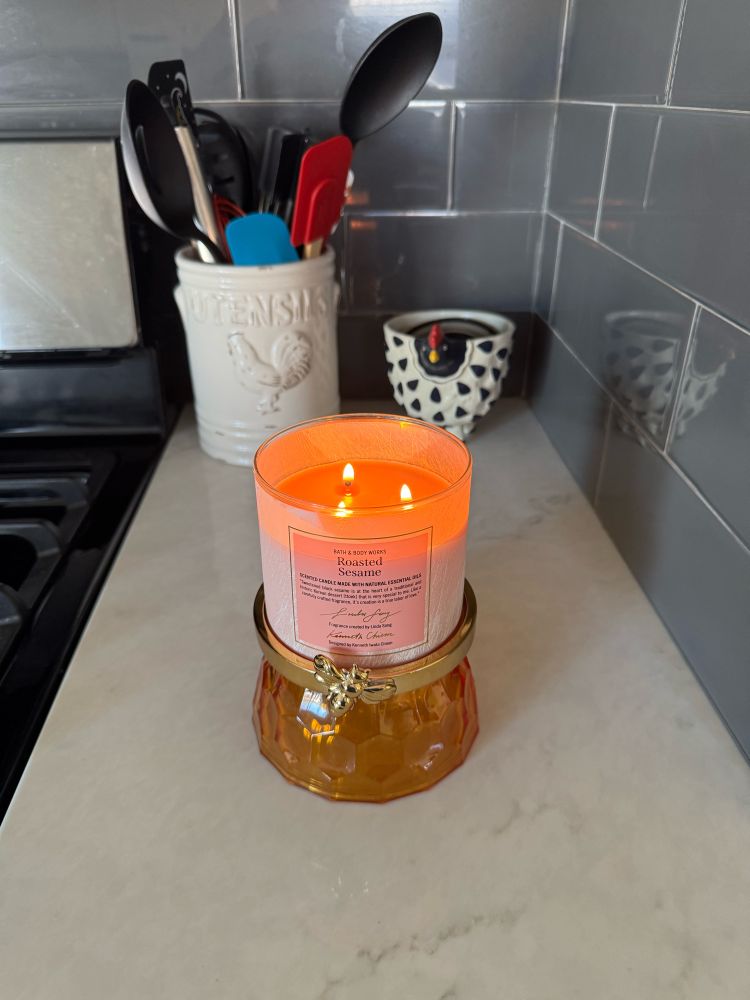 A lit candle in an ornate container sits on a marble countertop, beside a utensil holder and a glimpse of kitchen tools.