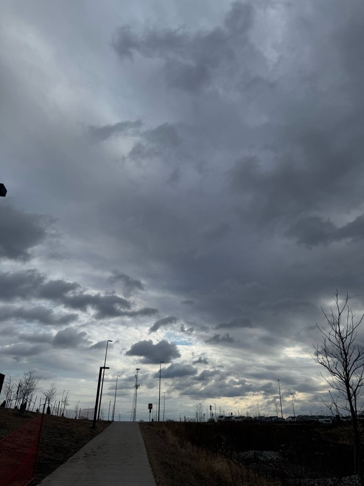 Dramatic gray clouds dominate the sky, with a glimpse of sunlight breaking through, alongside sparse trees and a pathway below.