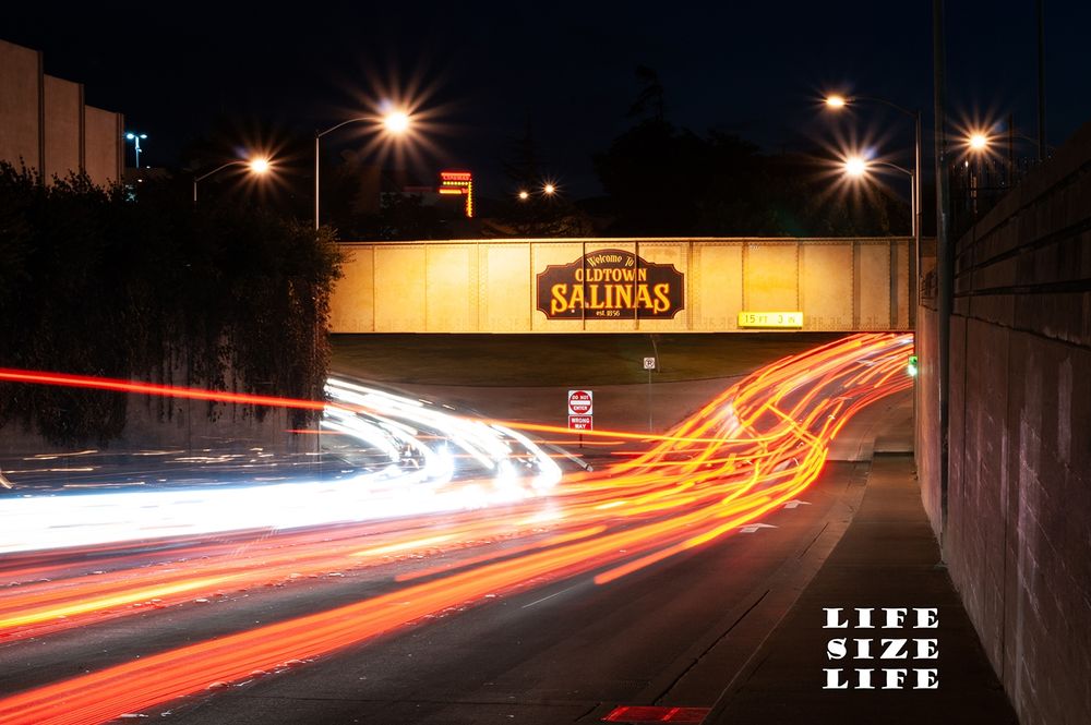 Streaks of light caused by taking a long exposure photo of vehicles driving under a bridge with "Welcome to Oldtown Salinas" painted on it. 