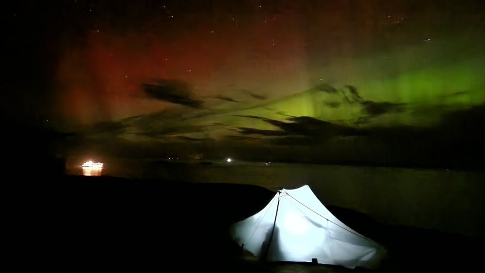 Photo of an illuminated tent at night. In the background is the Northern Lights (green and red patterns) against some black clouds. To the left is an illuminated ferry crossing the sea between the Isle of Skye and the Outer Hebrides.