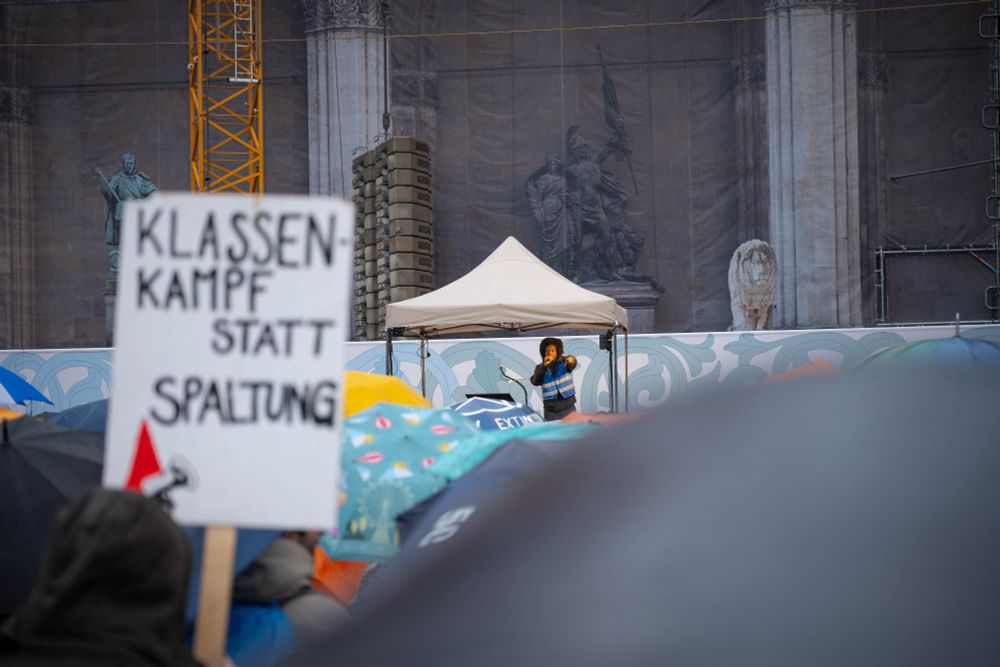 Jireh auf der Bühne, im Vordergrund Schirme und ein Schild "Klassenkampf statt Spaltung"