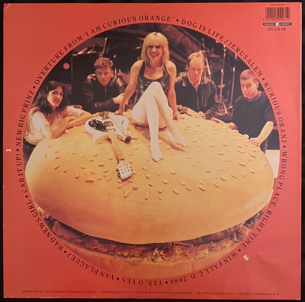 Back cover of the 1988 vinyl LP by The Fall, I Am Kurious Oranj. Features the track list and a photo of the members of The Fall except singer Mark E. Smith: Marcia Schofield, Craig Scanlon, Brix Smith, Steve Hanley, and Simon Wolstencroft. The band are arrayed around a giant hamburger bun, with Brix Smith sitting on top smiling. 